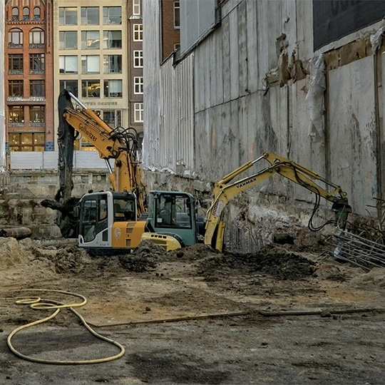 Two earthworks machines working within a large hole in an urban construction site.